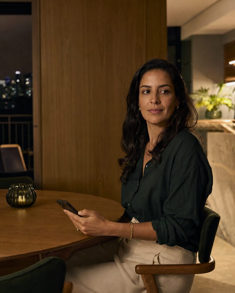 Mulher adulta observa a noite pela janela enquanto segura o celular